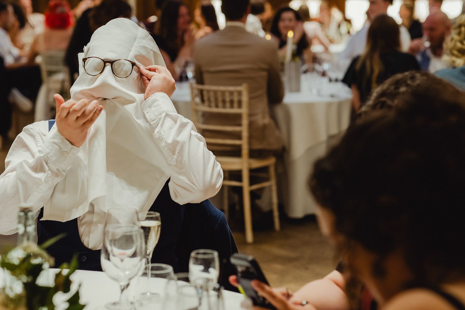 Wedding guests react emotionally during speeches at an indoor reception venue with white chairs and table settings.