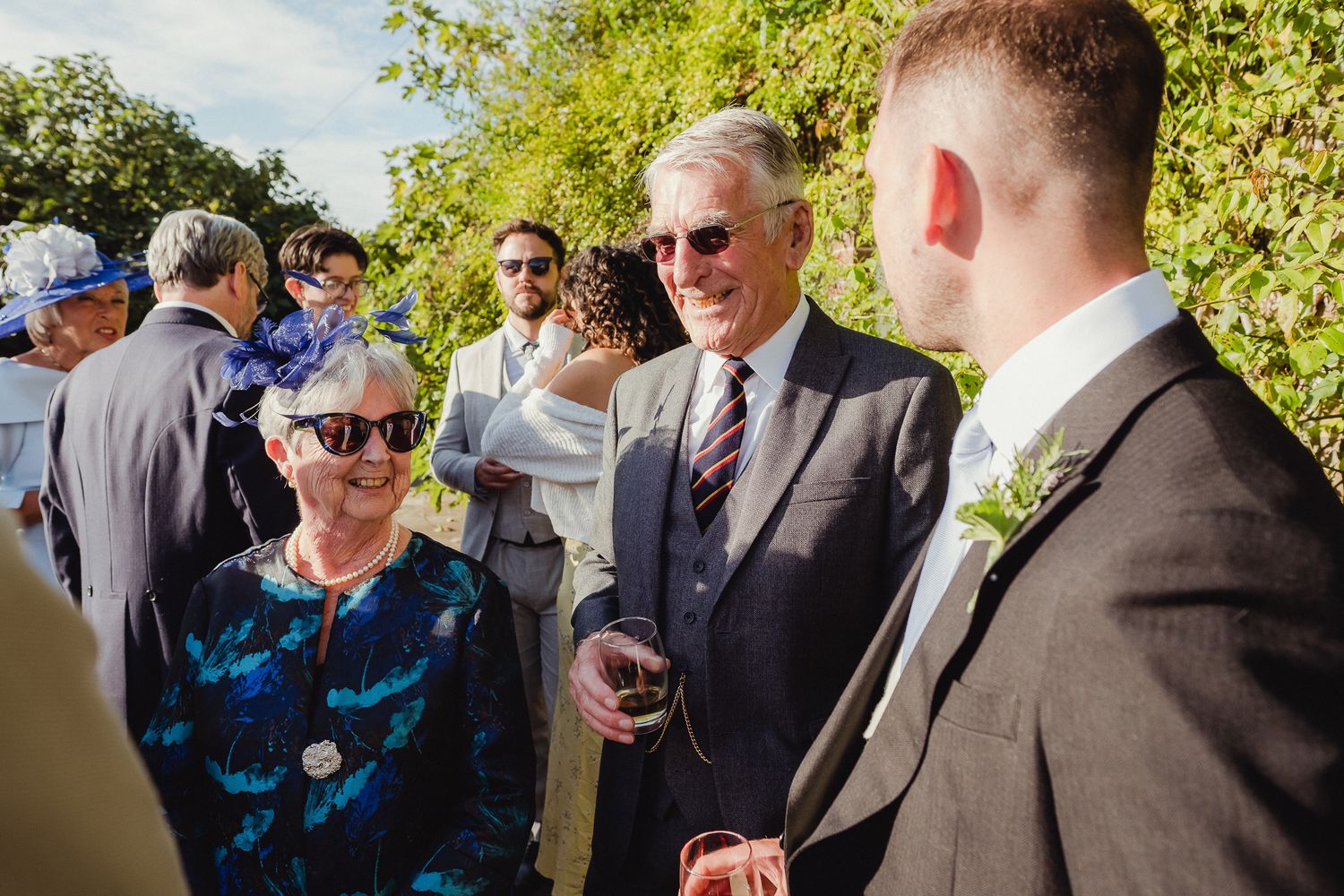 A group of people in formal attire gather outdoors in a sunny garden setting for a social event.