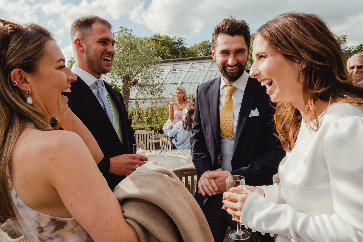 Wedding party laughs together during outdoor celebration in bright natural lighting.