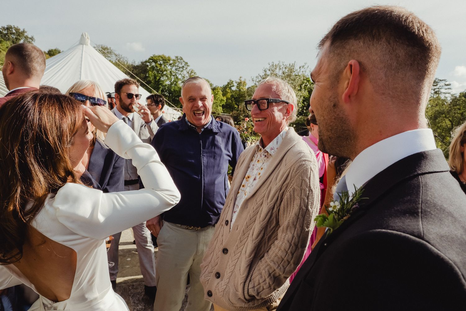 Wedding guests chatting and mingling outdoors at a sunny daytime reception with a white tent in the background.