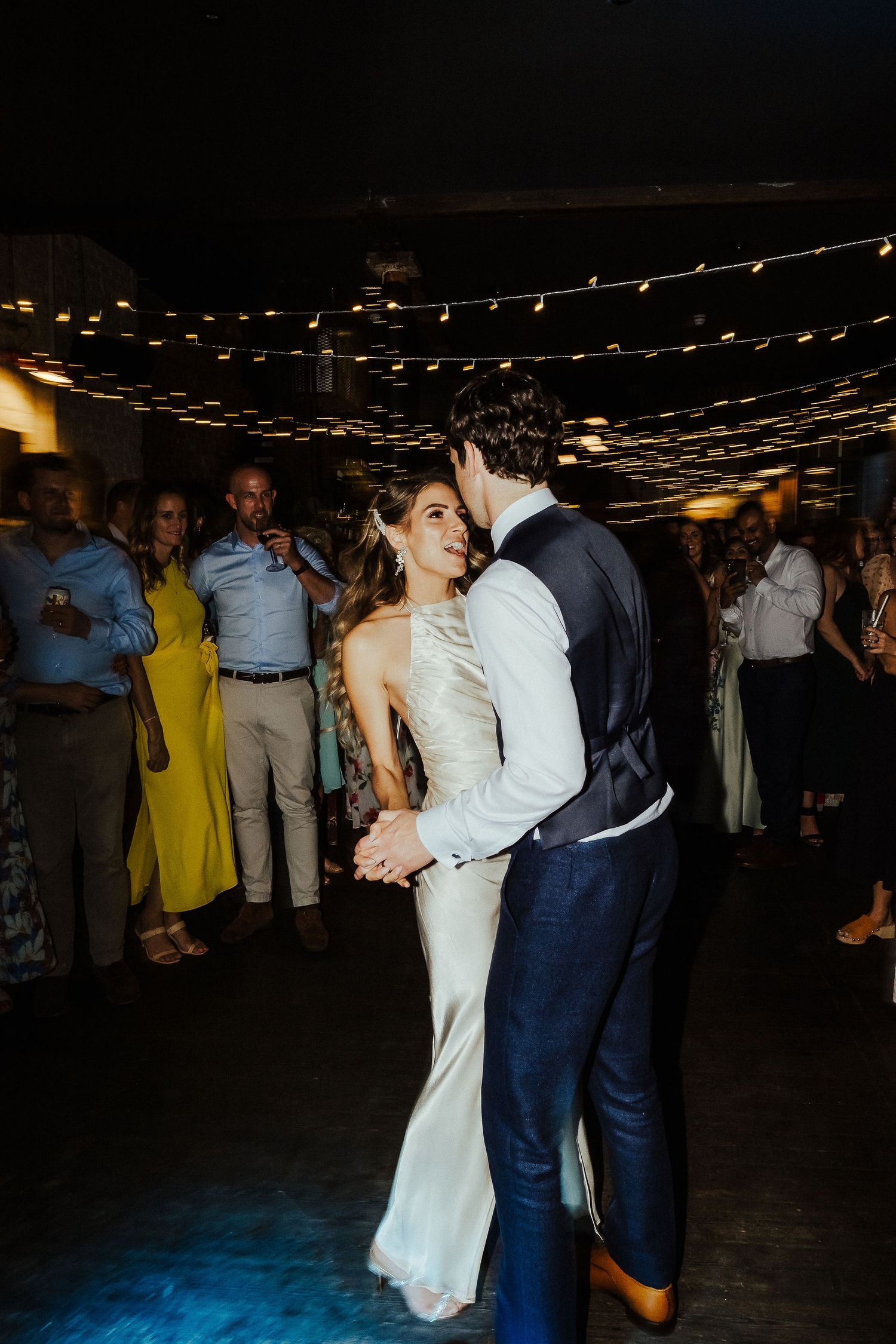 Couples slow dance together on a dimly lit dance floor under twinkling string lights at an evening reception.