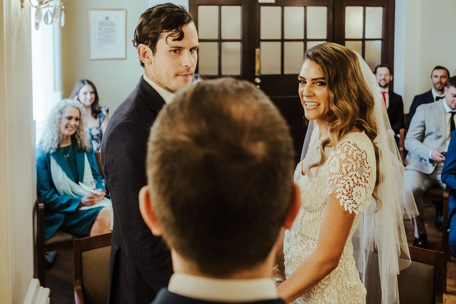 Wedding ceremony guests gather in an elegant indoor venue with natural light streaming through windows.