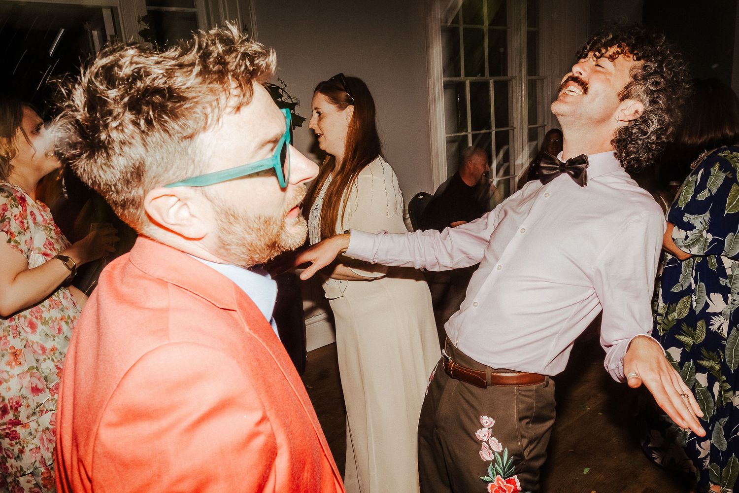 Wedding guests dance joyfully at a lively party, one man in teal sunglasses and orange blazer, another in a bow tie and suspenders.