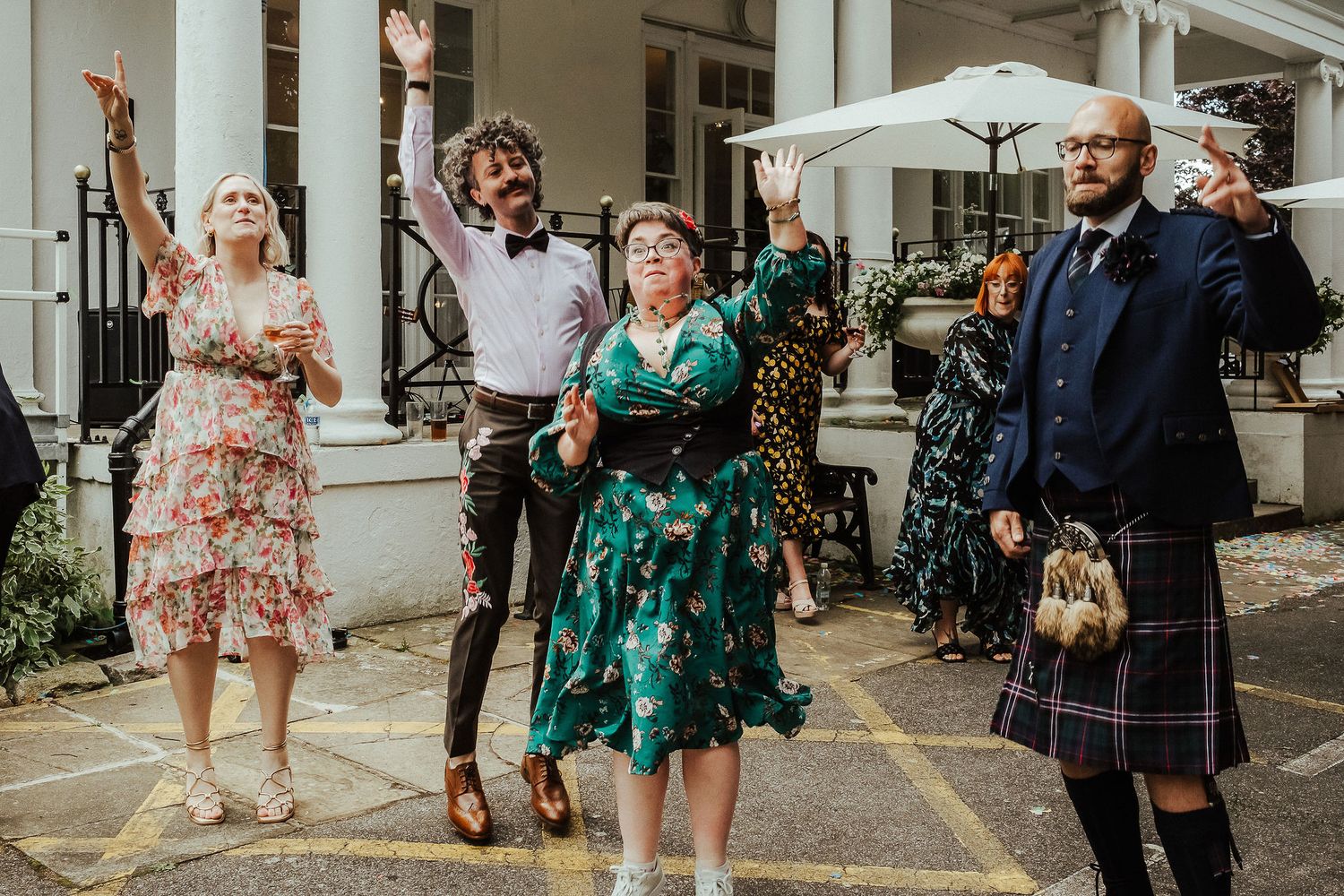 Wedding guests in colorful outfits, including a man in a kilt, cheer with raised arms on a sunny outdoor patio.