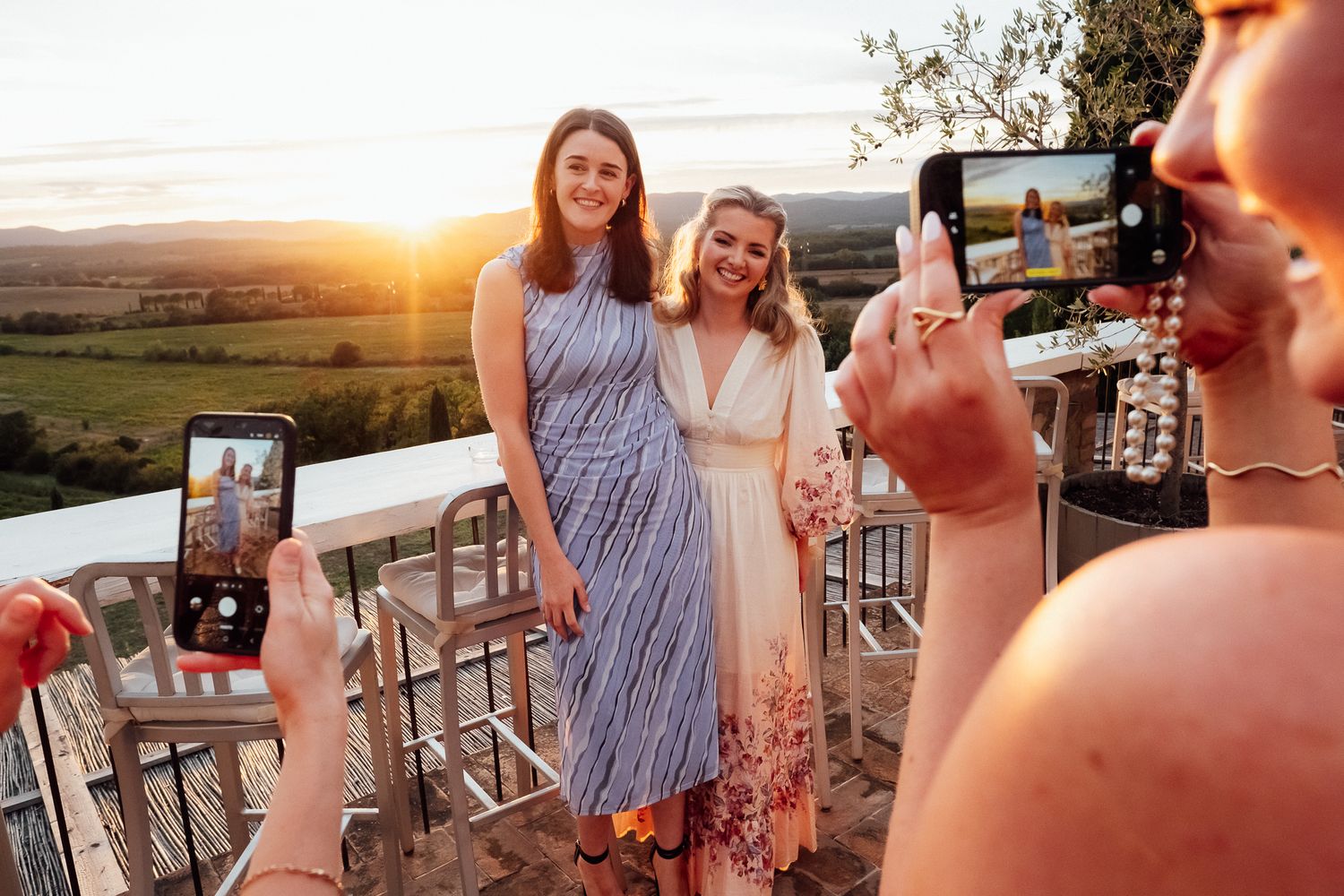 Two smiling women pose on a rooftop terrace at sunset while friends photograph them with smartphones, overlooking rolling countryside.
