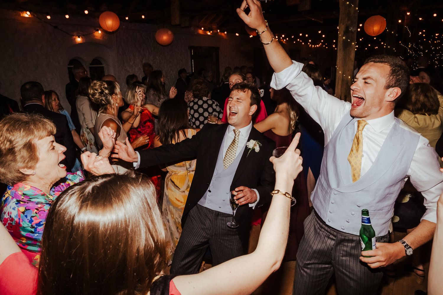 Wedding guests dancing and celebrating together during a lively reception party with festive lighting.