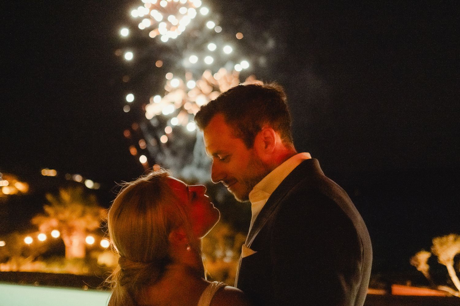 Romantic couple embraces under twinkling string lights at outdoor wedding reception during evening celebration.