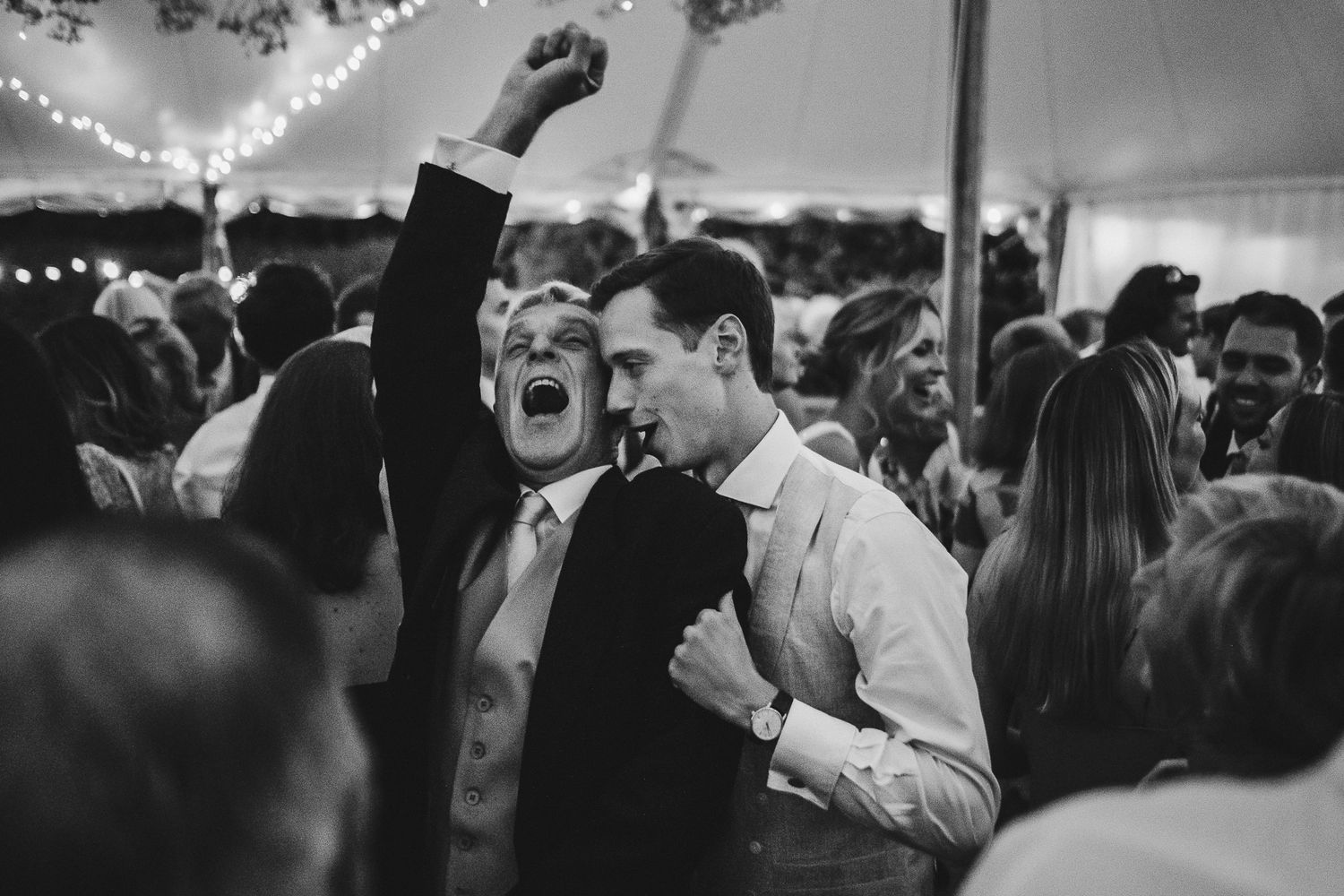 Guests celebrating with enthusiastic dancing and raised arms at a wedding reception under string lights.