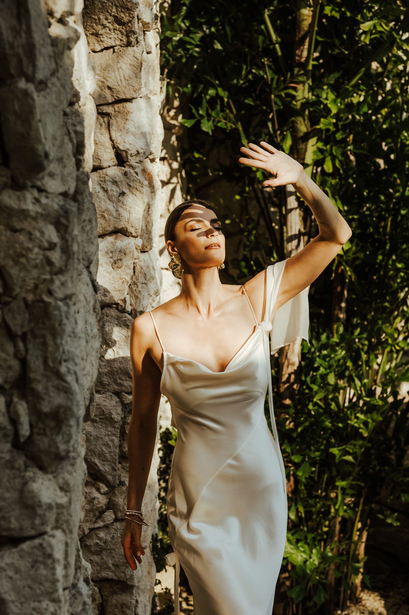 Person in white silk dress poses gracefully against stone pillars in a sunlit garden setting.