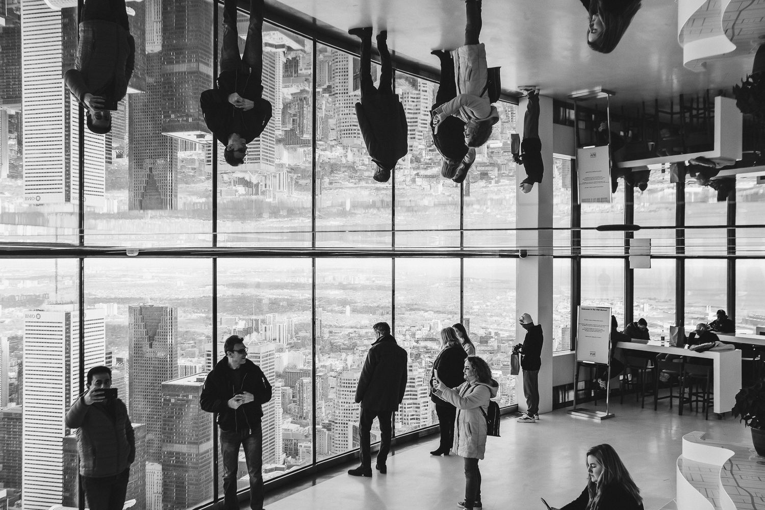 Black and white photo of people walking through an office space with hanging lamps and large windows.
