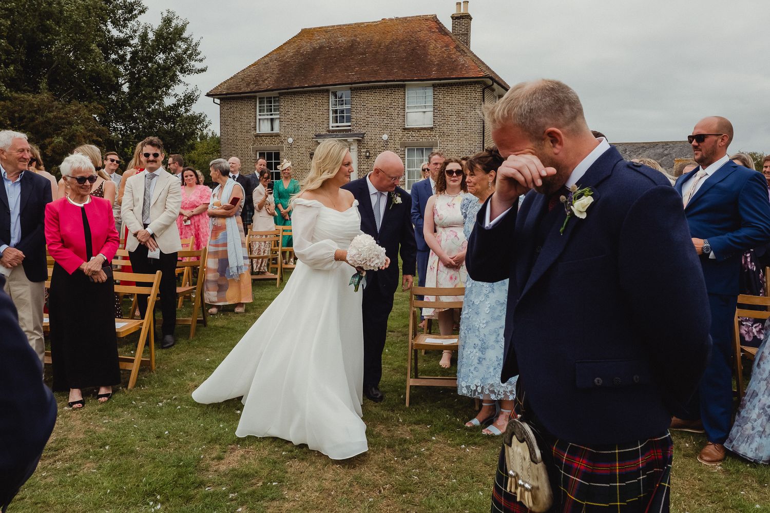 Wedding guests gather outdoors at a rustic countryside ceremony with a stone house in the background.