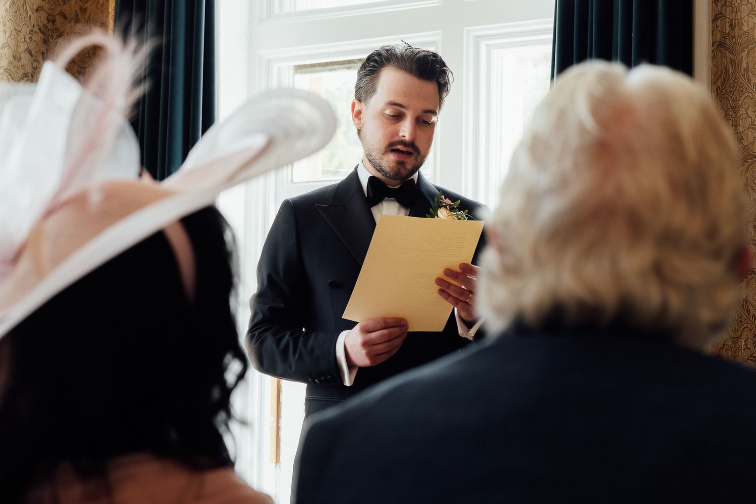 A groom in a tuxedo reads vows from a yellow card during a wedding ceremony.