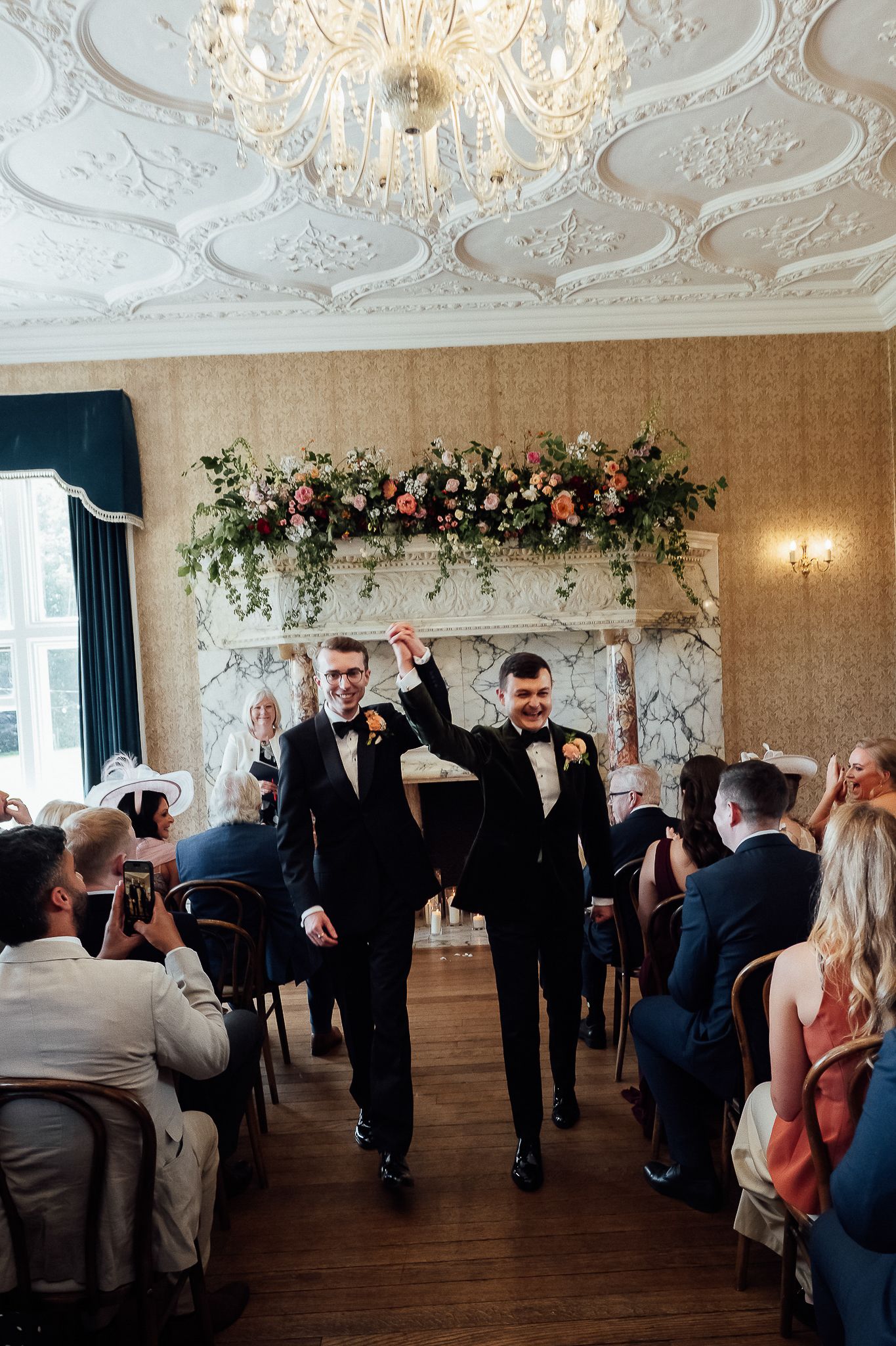 Two grooms celebrate their wedding ceremony in an elegant ballroom with floral arrangements and chandelier.