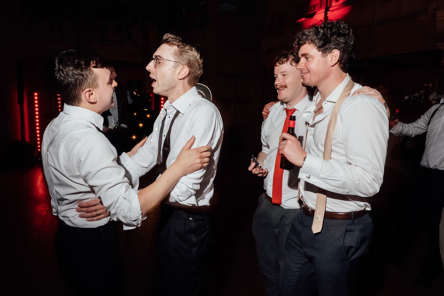 Four young men in white shirts and suspenders socializing on a dimly lit wedding reception dance floor.