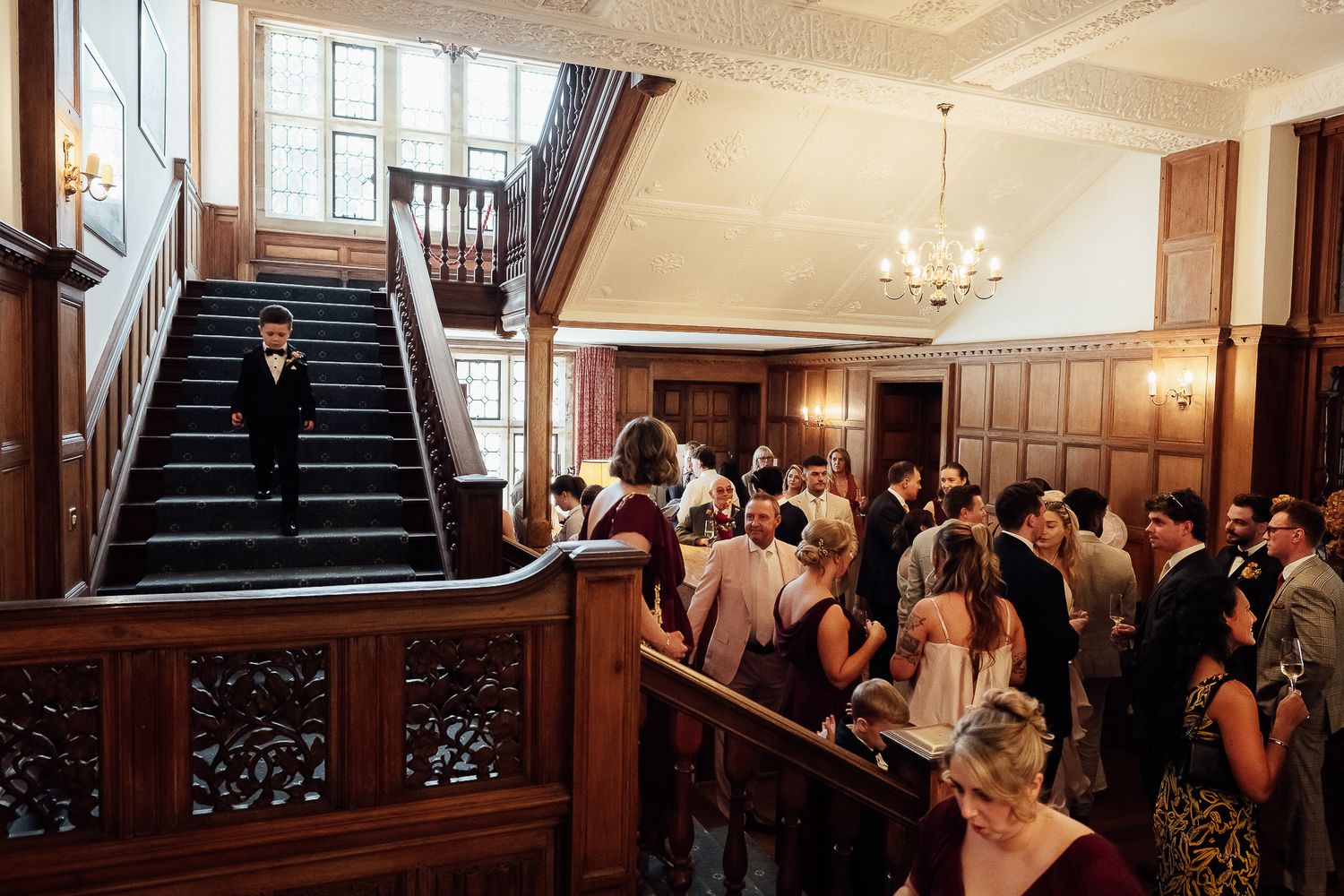 Elegant wedding reception in a grand wood-paneled hall with ornate staircase, chandelier, and formally dressed guests mingling.