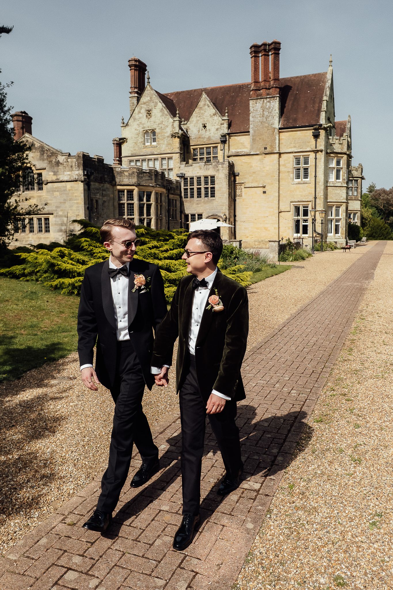 Two grooms in black tuxedos walk hand in hand outside a grand English manor on their wedding day.