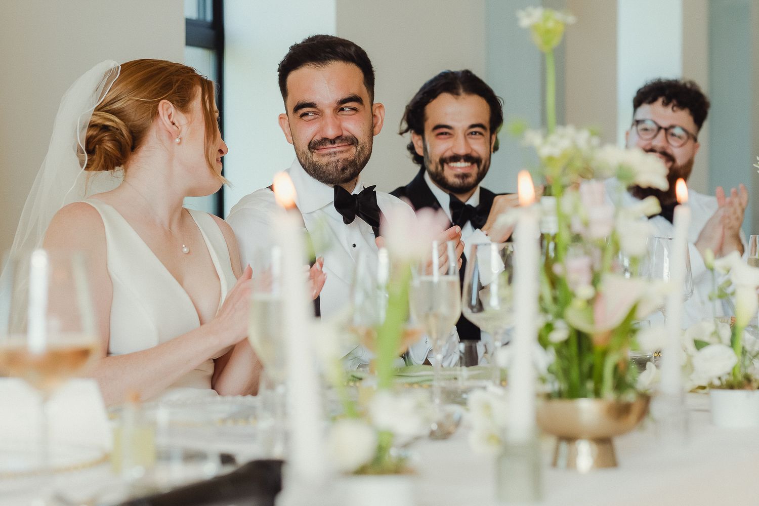 Wedding reception guests smile and converse at an elegant table decorated with white floral centerpieces and wine glasses.