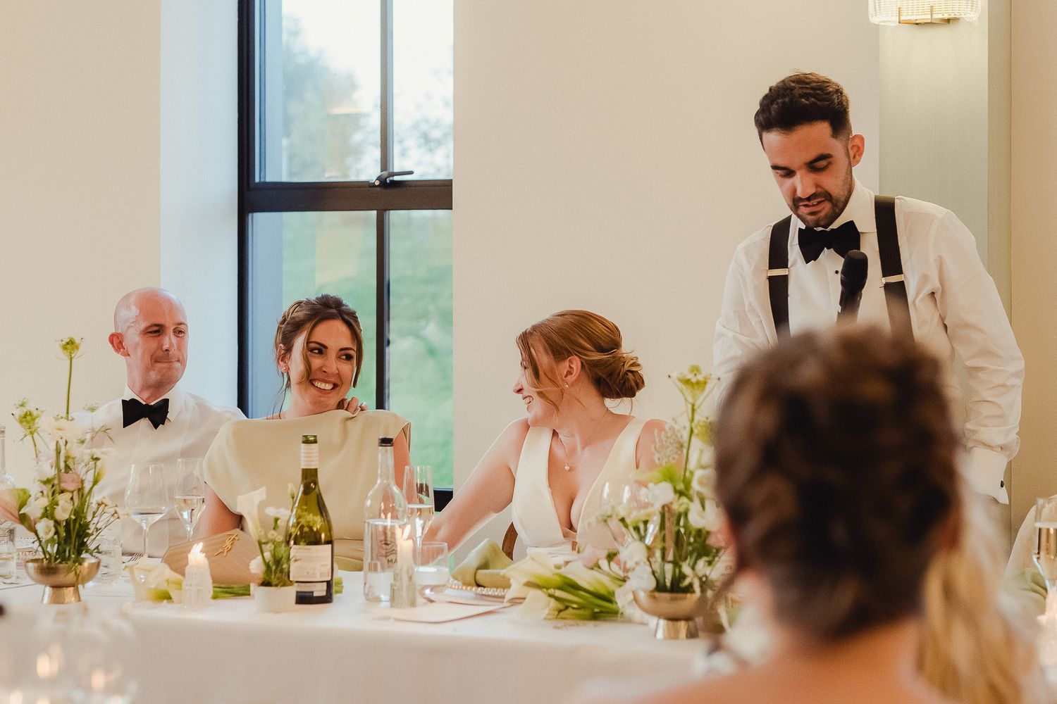 Wedding guests seated at a long dining table during a reception with floral centerpieces and wine glasses.