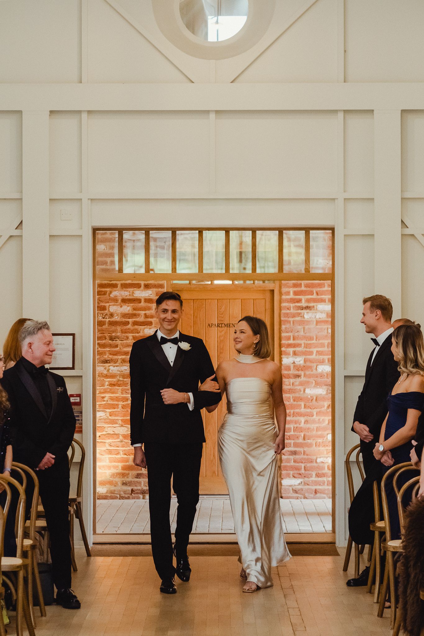 A couple walks down the aisle at their wedding ceremony in an elegant venue with exposed brick and wooden chairs.