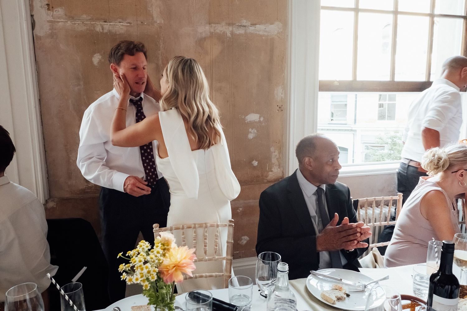 Wedding guests chat and laugh at a reception table with fresh flowers, wine and place settings in a sunlit venue.