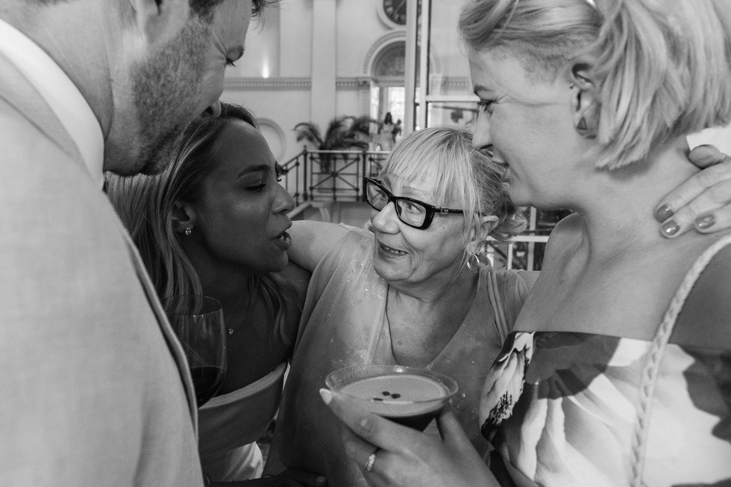 Group of smiling friends embracing in a close huddle at a celebration, one woman holding a cocktail in a black and white photo.