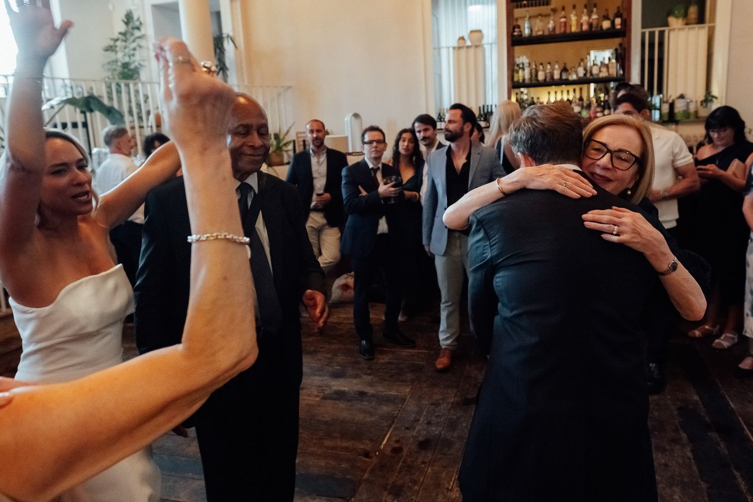 Wedding guests embrace and dance in an elegant room with a bar shelf in the background as a bride in a white dress raises her arm.