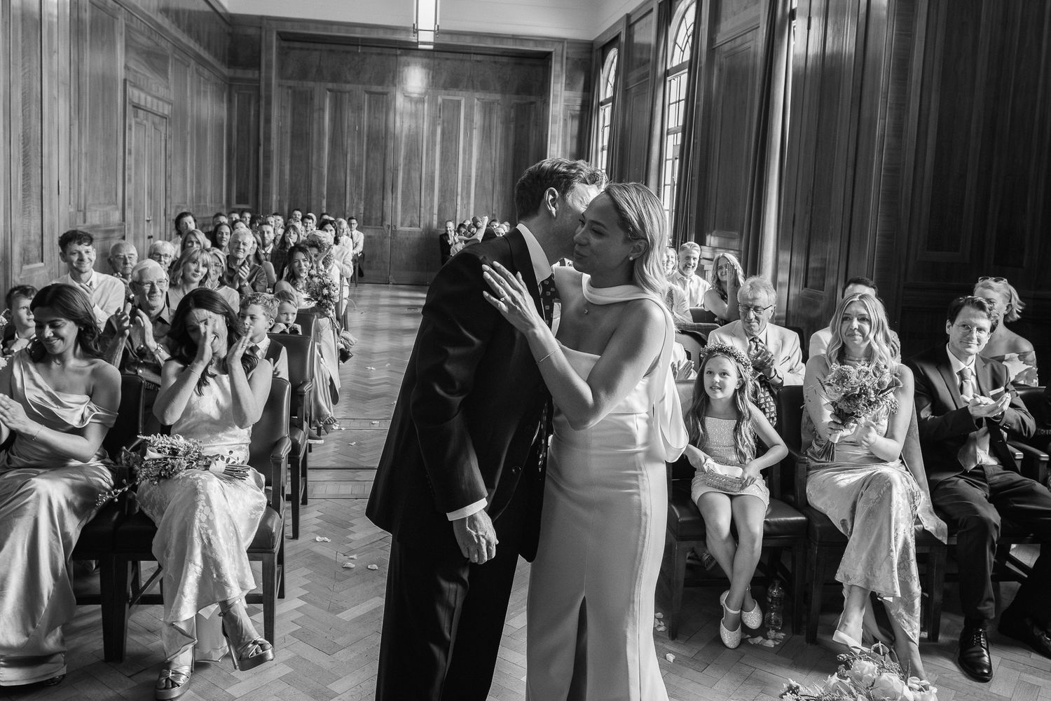 Bride and groom share a kiss during their wedding ceremony in a wood-paneled room as seated guests look on and smile.