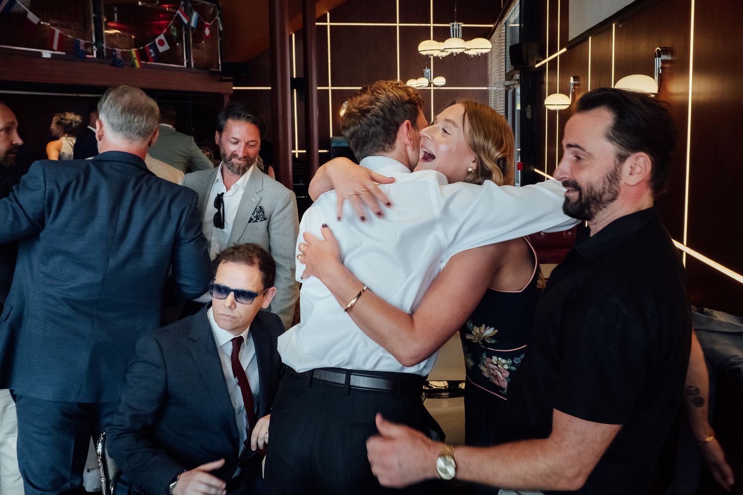 Two people share a warm embrace in a lively indoor gathering while a bearded man in black and guests in suits look on.