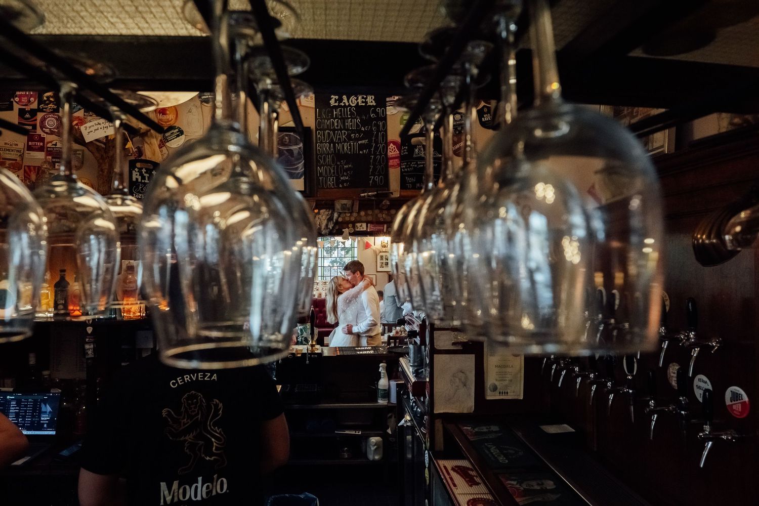 Rows of wine glasses hang upside down above a rustic bar while a bartender works near beer taps and a chalkboard menu in a cozy pub.