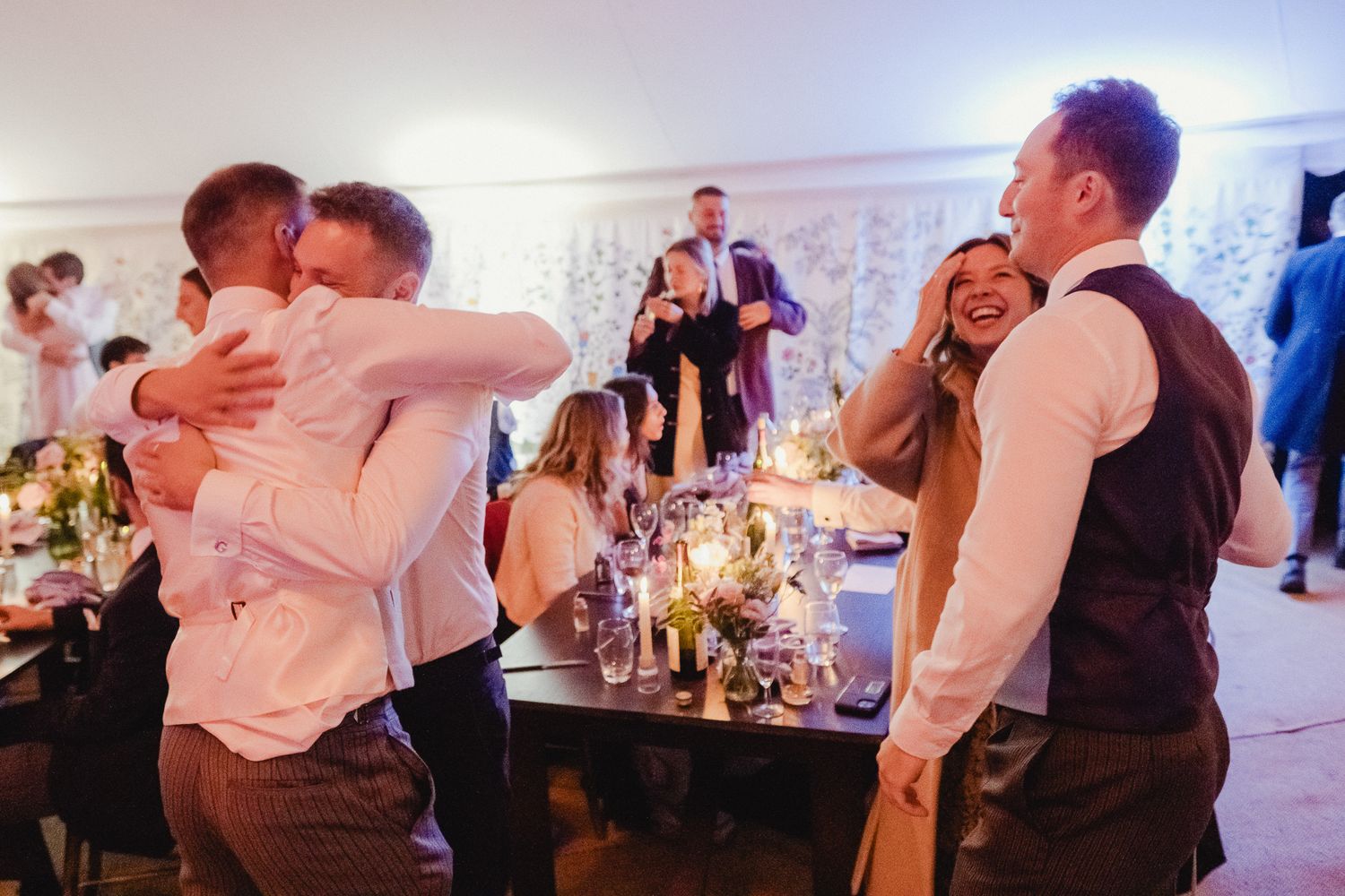 Wedding guests dance and embrace on the dance floor during an elegant reception celebration in a white-decorated venue.