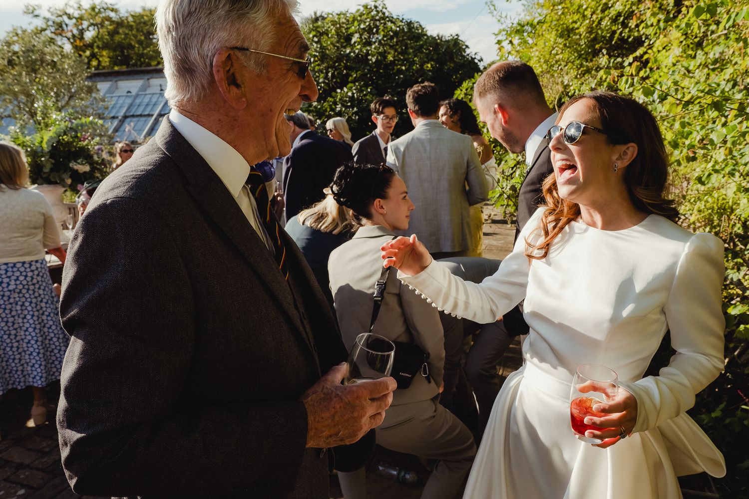 Wedding guests gather outdoors in a garden setting for a celebration with drinks and laughter.