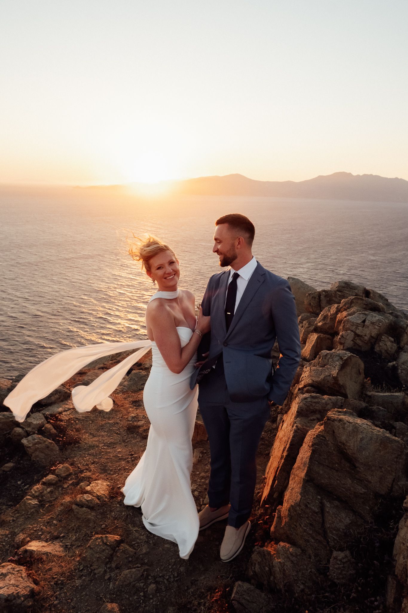 Bride and groom embrace on rocky cliffside at golden sunset with ocean and mountains in background.