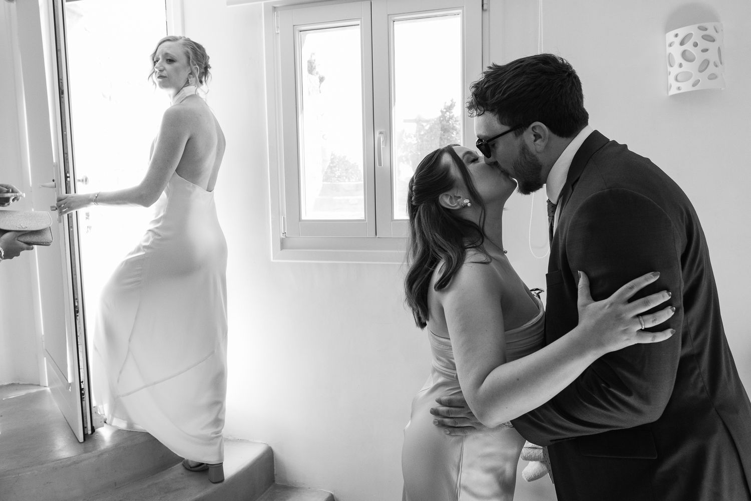 Black and white wedding photo of couple kissing while bride in elegant gown stands in doorway background.
