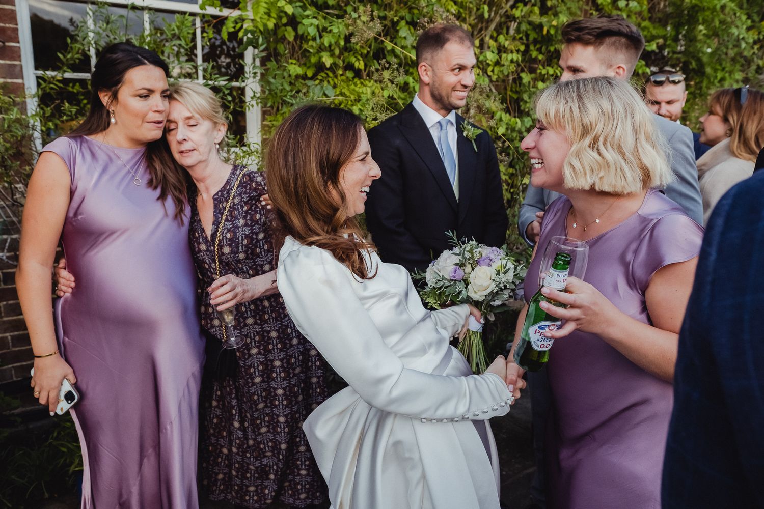 Wedding party members in purple dresses and formal attire share laughter and drinks during an outdoor celebration.