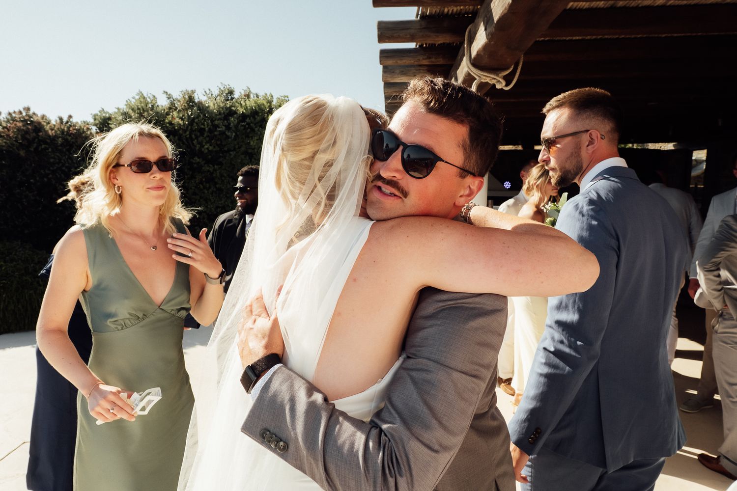 Bride and groom sharing a warm hug at outdoor wedding reception with guests in elegant attire nearby.