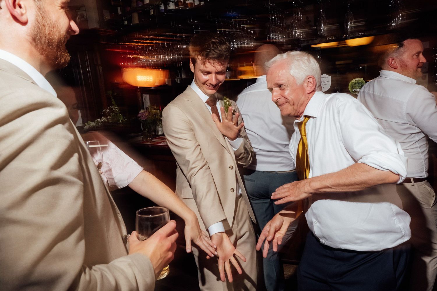 Smiling men in suits and shirts dance and celebrate together at a lively indoor party, one holding a drink in a warmly lit bar.