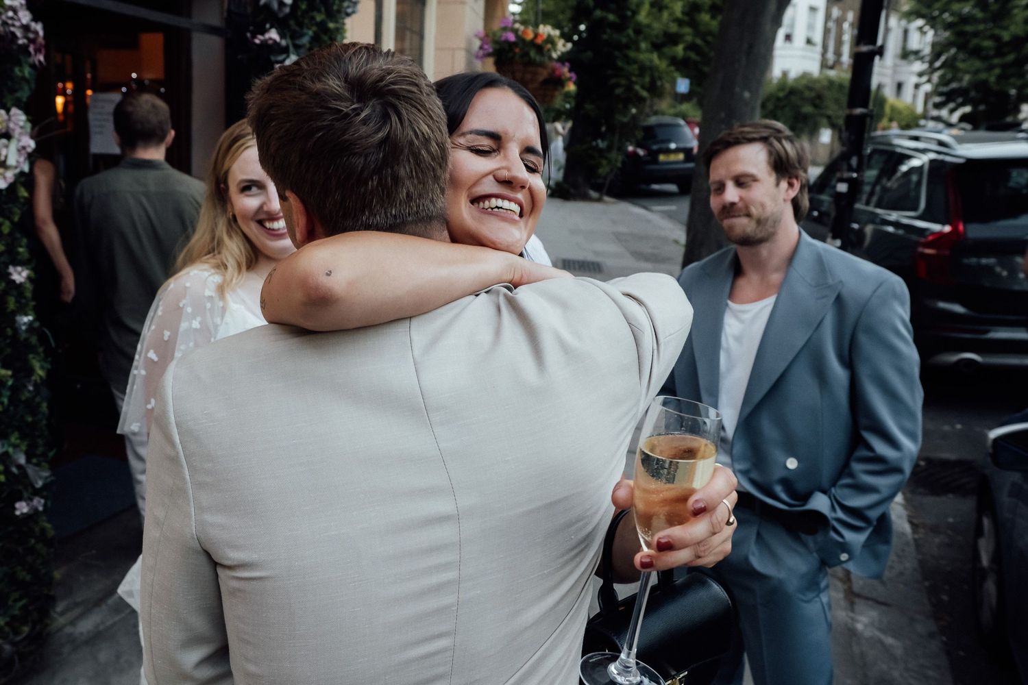 A smiling woman hugs a man in a light suit while holding a champagne glass at an outdoor celebration, with guests chatting nearby.