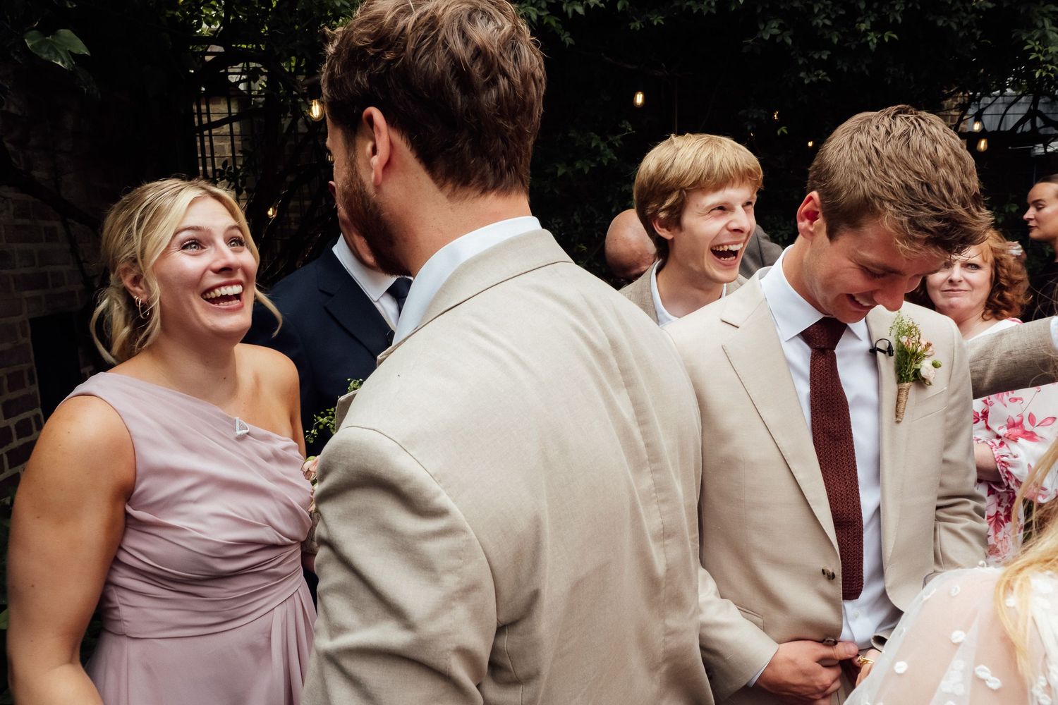 Guests laugh and celebrate at a wedding as a groom in a tan suit with a floral boutonniere stands among smiling people in formal attire.