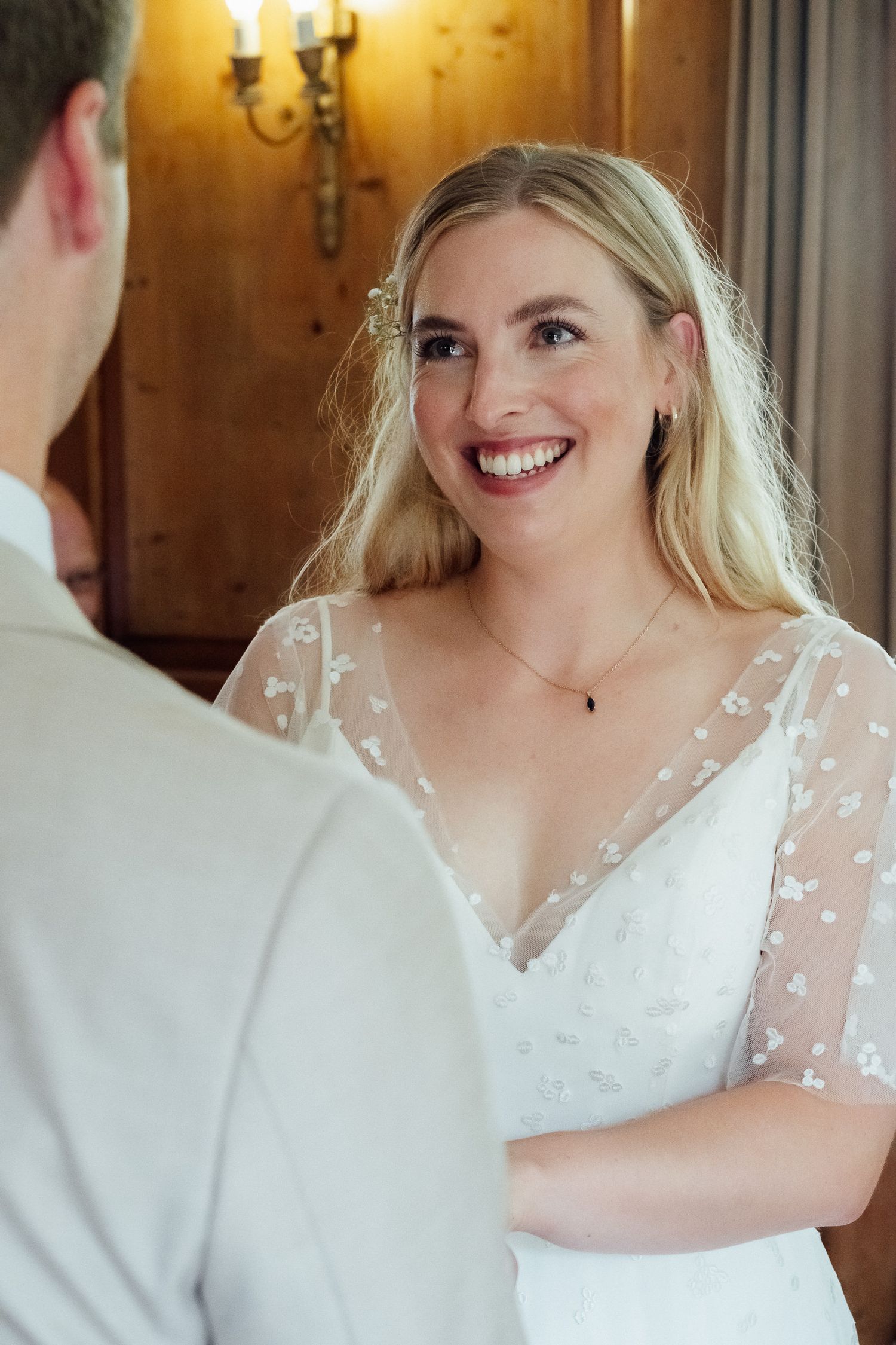 Smiling bride with long blonde hair wears a white floral lace wedding dress while facing her groom in a warm wood-paneled room.