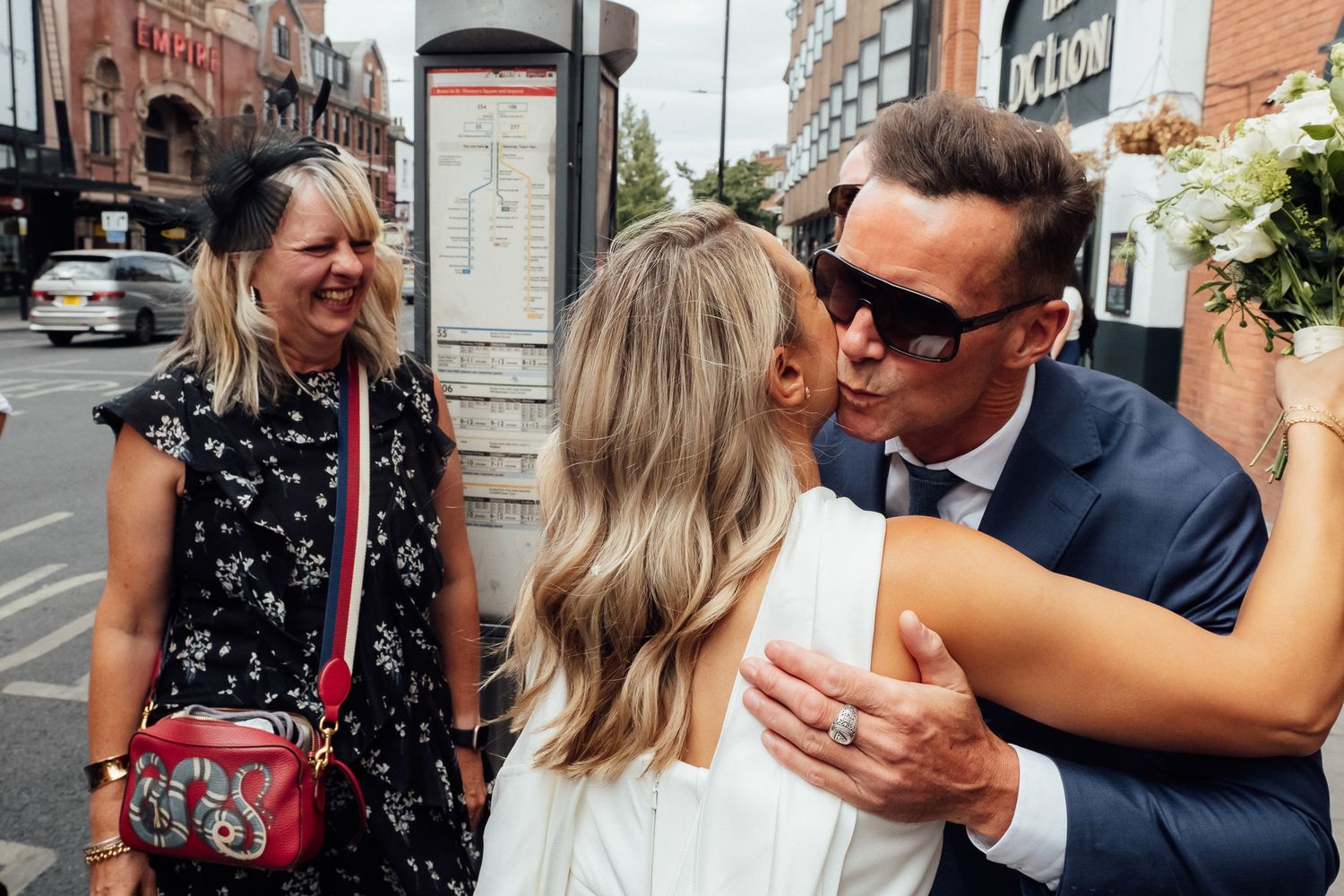 Bride in a white dress embracing a smiling man in sunglasses and a navy suit while a woman with a red crossbody bag looks on.