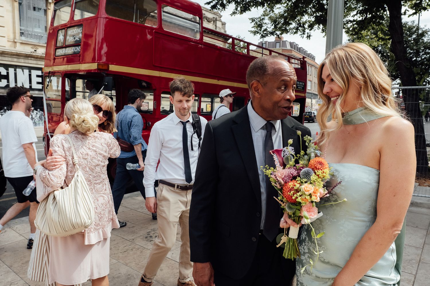 Wedding guests in formal attire gather on a city street near a red double-decker bus as a woman in a pale dress holds a colorful bouquet.