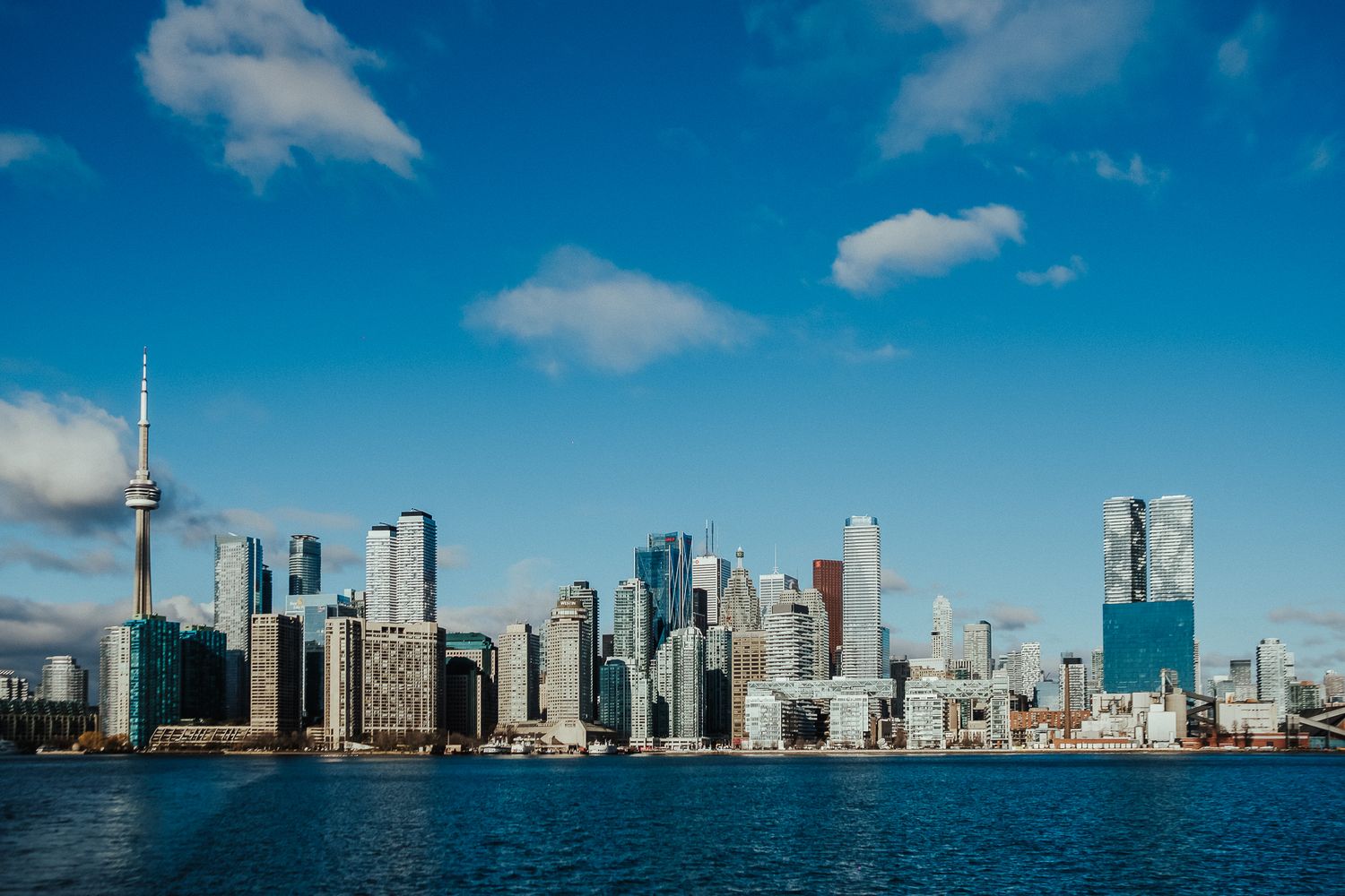 Panoramic skyline view of Toronto's downtown waterfront with modern skyscrapers against a bright blue sky.
