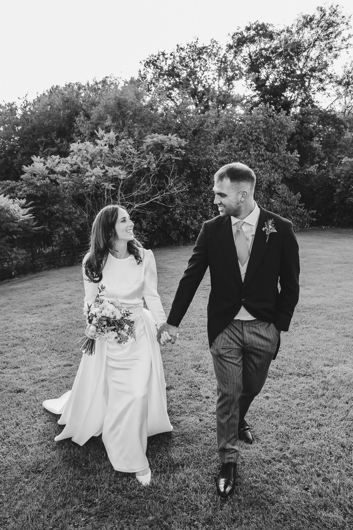 A couple in wedding attire playfully walks and dances together in a grassy outdoor field.