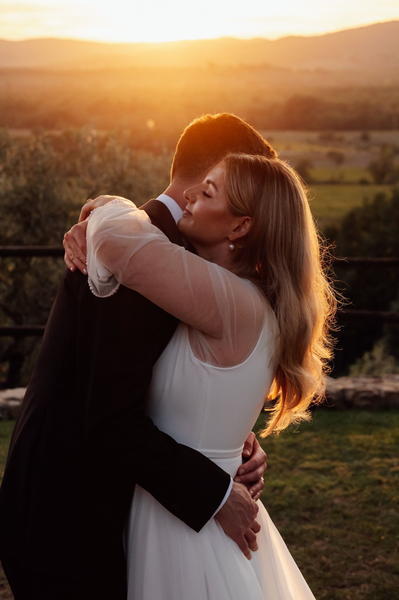 Bride in a white tulle gown embraces her groom in a dark suit during a golden sunset overlooking rolling countryside hills.
