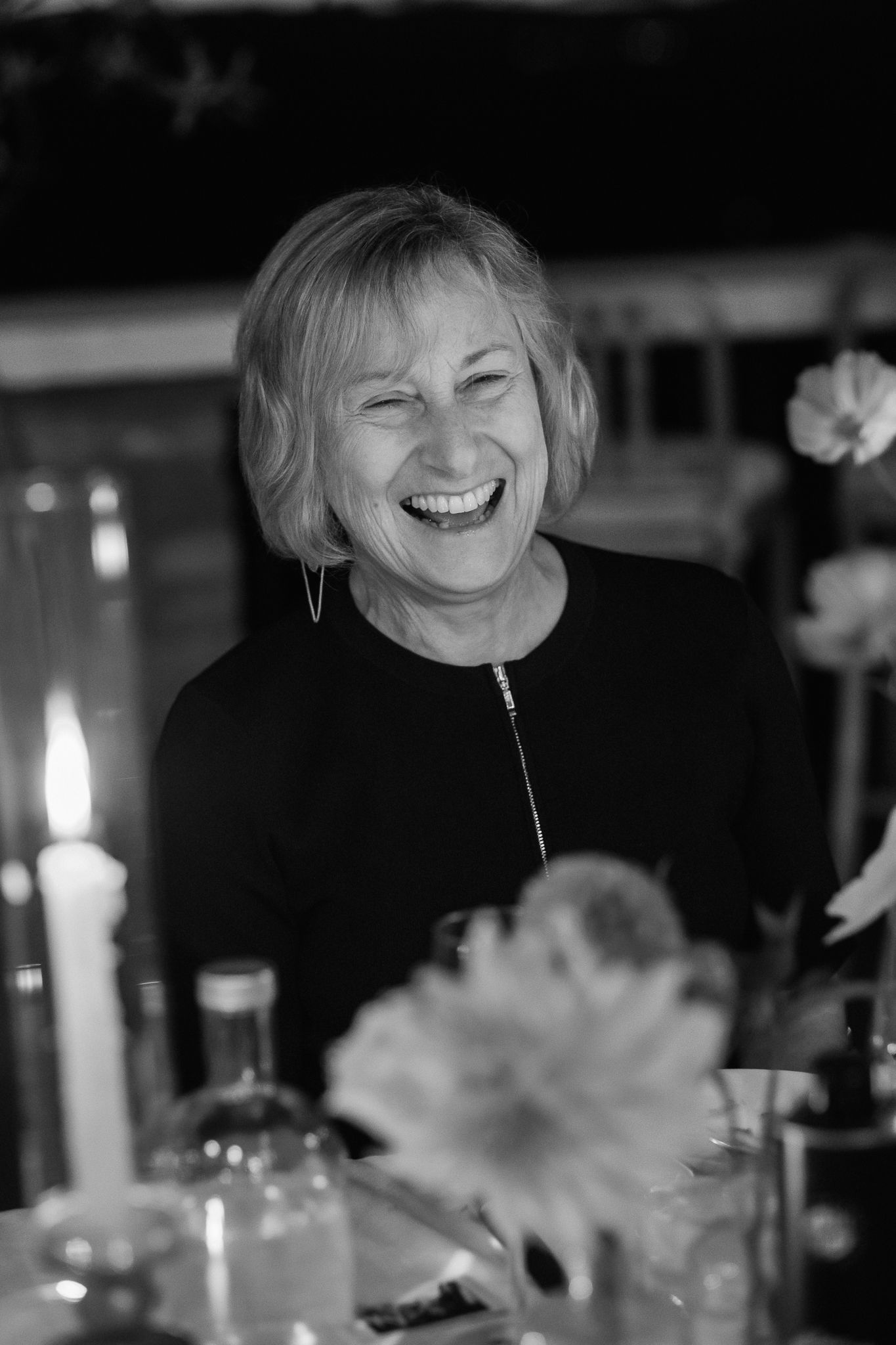 Black and white photo of a laughing woman with short hair seated at a candlelit table with flowers and glass bottles.