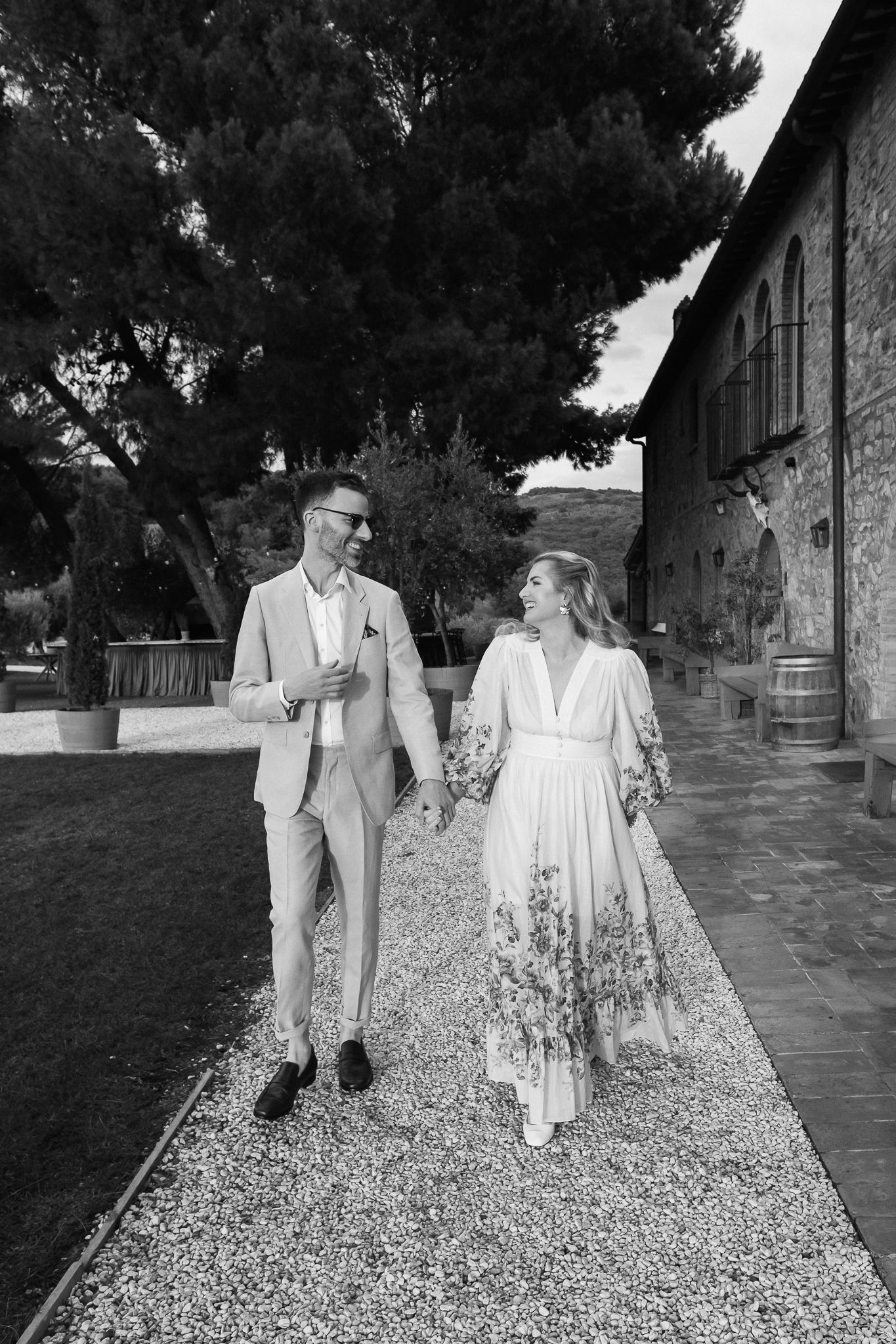 Black and white photo of a smiling couple holding hands and walking on a gravel path beside a stone building, perhaps on a wedding day.