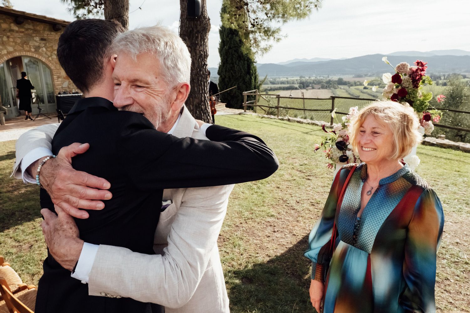 Older man in a white shirt hugs a man in a black suit at an outdoor wedding while a smiling woman holds flowers, with hills behind.