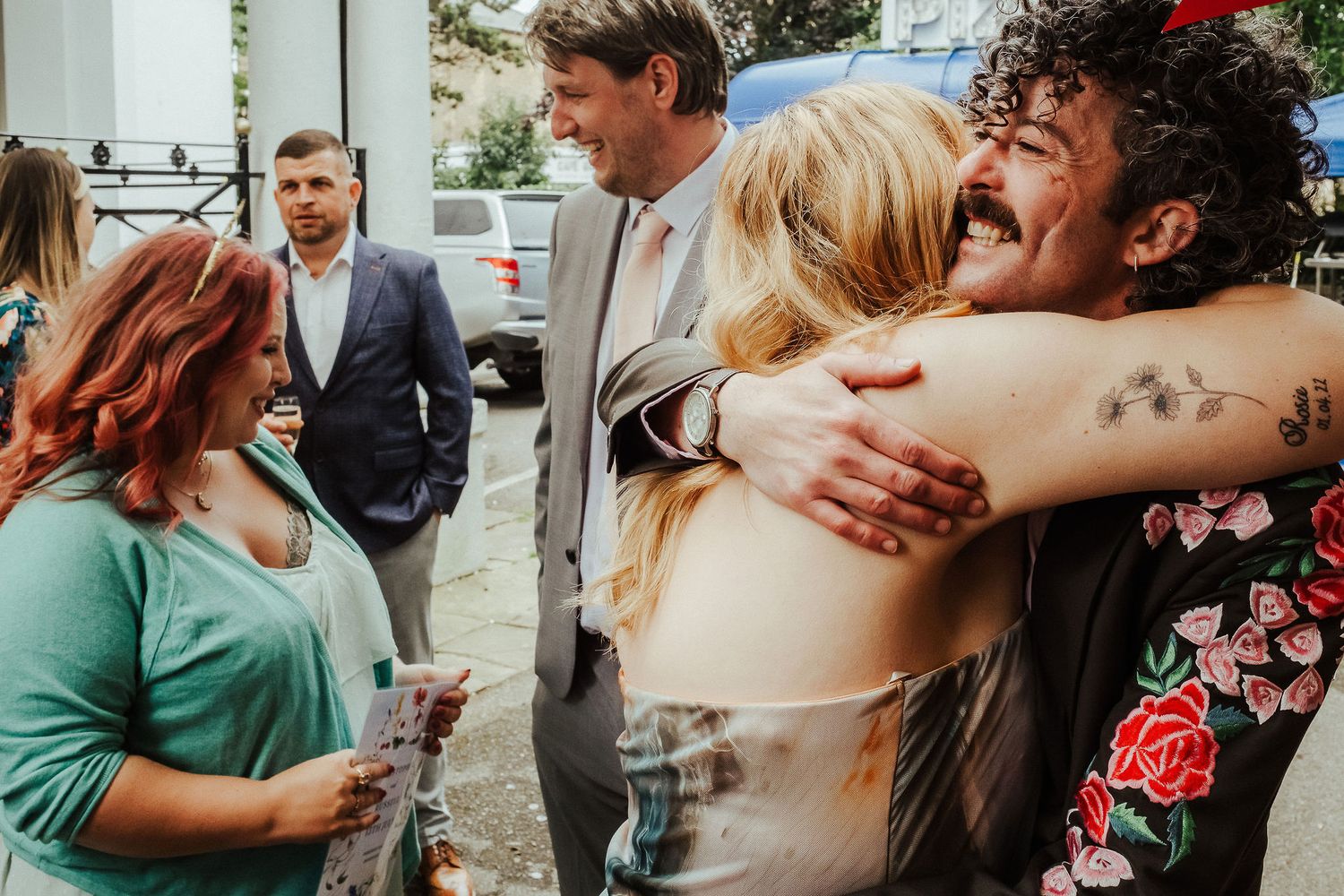 Wedding guests embrace warmly outdoors as a curly-haired man in a floral shirt hugs a blonde woman while others smile nearby.