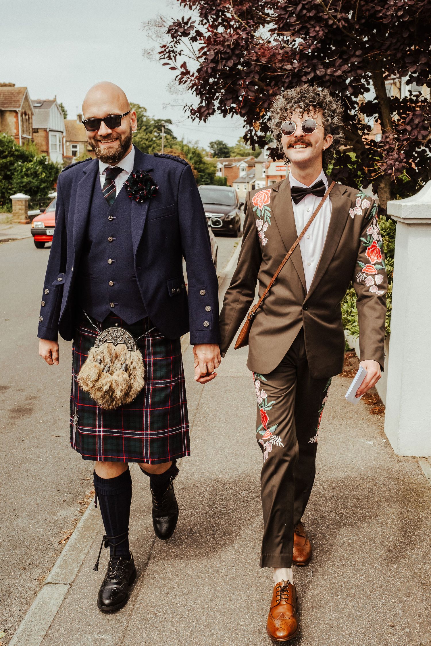 Two men holding hands walk down a sunny residential street, one in a navy Scottish kilt outfit and the other in an embroidered floral suit.