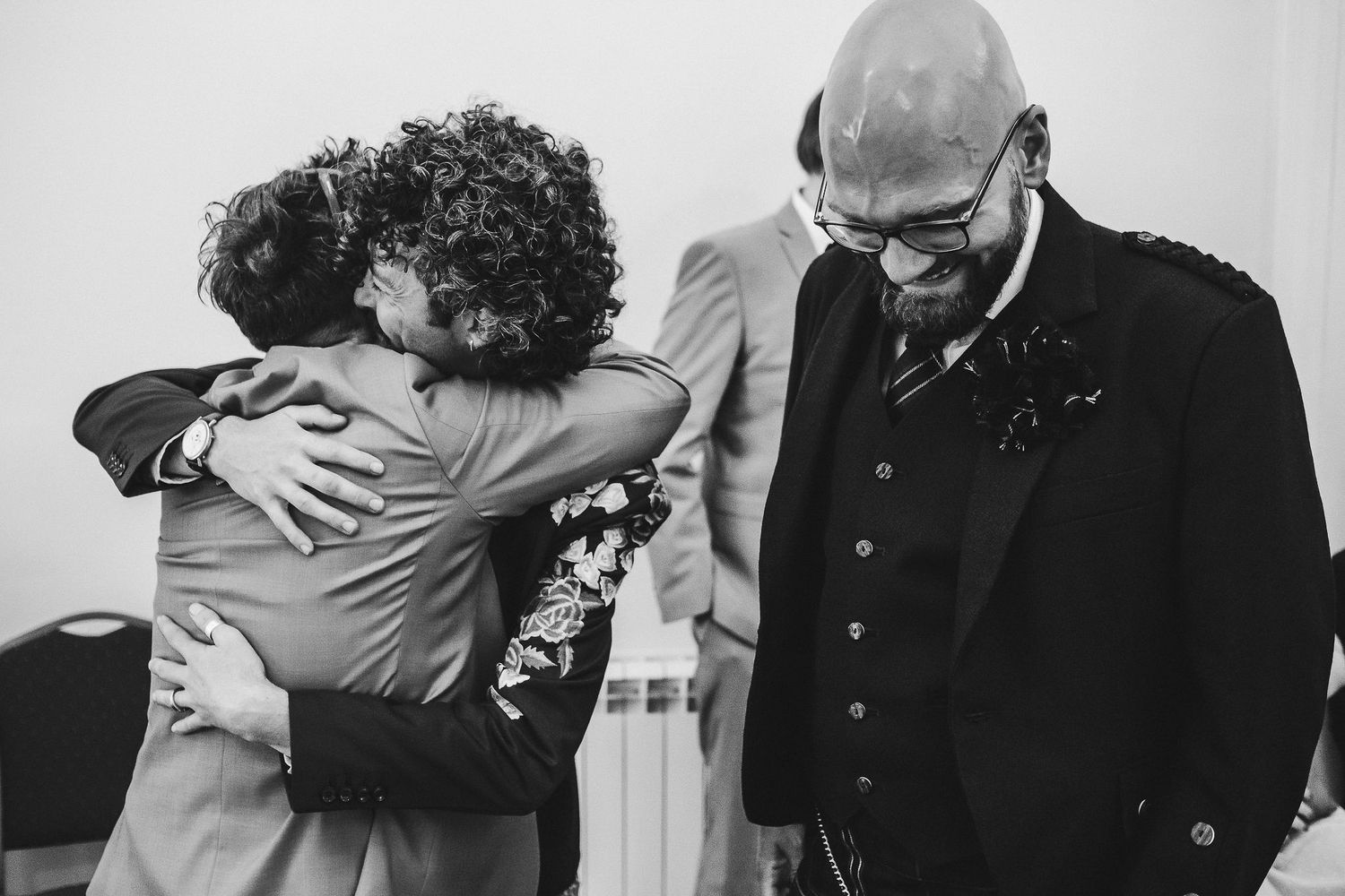 Black and white photo of two men embracing warmly at a wedding while a bald, bearded man in glasses and a boutonniere smiles nearby.